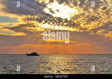 Dramatischer Sonnenuntergang über dem Meer mit Sonnenstrahlen, die durch Wolken dringen, und einer Yacht, die ruhig am Horizont schwimmt. Stockfoto