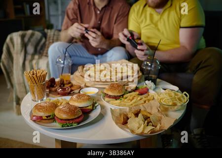 Zwei Freunde sitzen und genießen eine lustige Gaming-Session mit einer großen Auswahl an leckerem Fast Food auf einem kleinen Tisch. Tisch gefüllt mit Burgern, Pommes und anderen Snacks Stockfoto