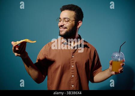 Lächelnder Mann mit Bart, Brille, Pizzablätter in der einen Hand und ein Getränk in der anderen, vor blauem Hintergrund. Hispanischer Mann genießt sein Essen Stockfoto