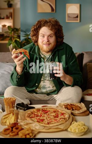 Mann lächelt und hält Pizzascheiben in der Hand, während er auf einer Couch mit verschiedenen Snacks und einem Getränk sitzt und ein Wochenende mit Freunden zu Hause genießt Stockfoto