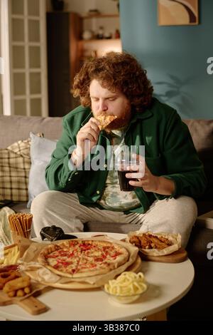 Mann, der auf der Couch sitzt und Pizza und Snacks genießt, während er Fernsehen sieht, umgeben von verschiedenen Speisen und einer gemütlichen Wohnzimmerumgebung Stockfoto