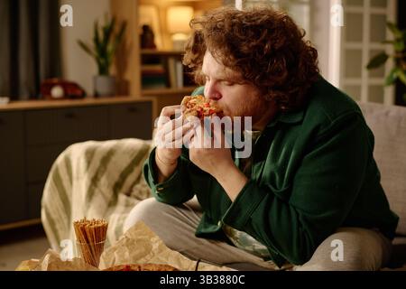 Bärtiger Mann, der auf einer Couch sitzt, genießt ein Stück Pizza in einem gemütlichen Wohnzimmer mit warmer Beleuchtung. Hintergrund enthält ein Sofa, dekorative Elemente und ein teilweise sichtbares Bücherregal Stockfoto