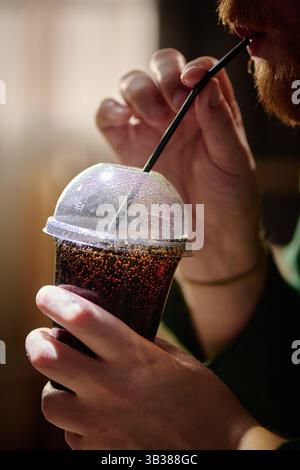 Nahaufnahme der Hand, die ein kaltes Getränk in einem Plastikbecher mit Strohhalm hält. Seitenprofil eines bärtigen Individuums, das erfrischende Getränke genießt. Stockfoto