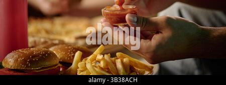 Eine Nahaufnahme einer Hand, die Pommes frites in eine kleine Glasschale Ketchup taucht, wobei Burger und weitere Pommes im Hintergrund zu sehen sind Stockfoto