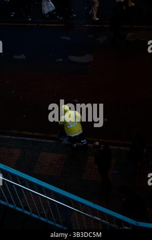 Ein Stadtarbeiter in Neonausrüstung fliegt unbemerkt, wenn die Menschenmassen am Abend auf einer dunklen Stadtstraße vorbeiziehen Stockfoto