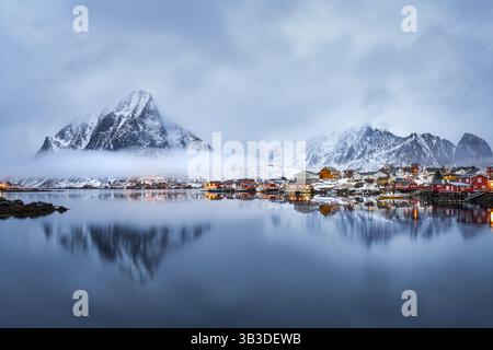 Winterblick über das Wasser in Richtung reine, Lofoten-Inseln, Norwegen, mit schneebedeckten Bergen und niedrigen Wolken. Stockfoto