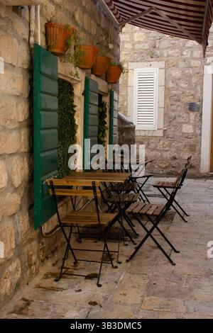 Gemütliches Café im Freien mit Holztischen und -Stühlen, vor alten Steinwänden und grünen Fensterläden. Stockfoto