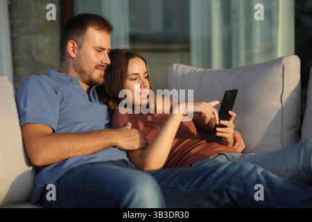 Entspanntes Paar bei Sonnenuntergang, das auf einer Couch auf einer Terrasse auf dem Handy sitzt Stockfoto