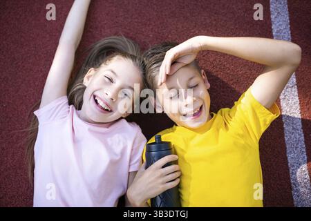 Glückliche zwei Kinder, ein Junge und ein Mädchen, liegen auf dem Rücken und lachen, während sie in die Kamera schauen, nachdem sie auf der Laufbahn des Stadions Joggingtraining gemacht haben. Sport Stockfoto