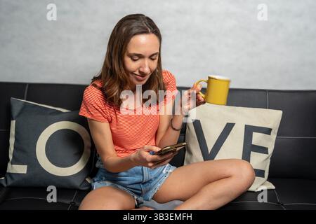 Ruhige junge Frau, die Kaffee genießt, während sie ihr Smartphone auf einem bequemen Sofa benutzt, um einen ruhigen Moment zu Hause zu genießen Stockfoto