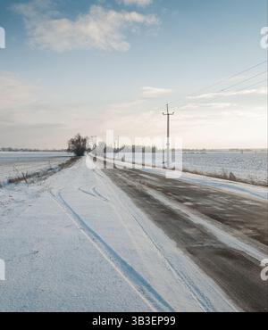 Landstraße im Winter. Landstraße in Winterfeldern an einem sonnigen Tag: Mit Eis bedeckter Asphalt, Freileitung rechts von der Straße. Stockfoto