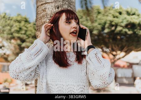 Frau in einem stilvollen weißen Pullover, die an einem sonnigen Tag draußen auf einem Smartphone spricht Stockfoto