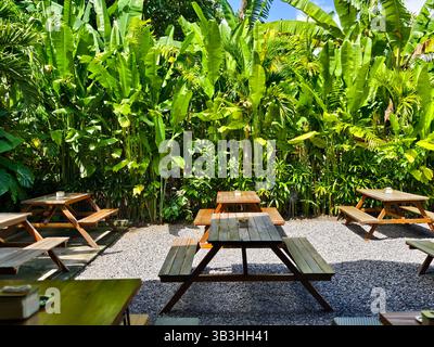 Sitzbereich im Café im Freien mit mehreren Picknicktischen aus Holz auf einem kiesbedeckten Boden, umgeben von üppigen tropischen Pflanzen Stockfoto