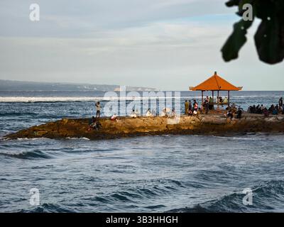 Sanur, Bali, Indonesien – 23. April 2025: Eine Gruppe von Menschen versammelt sich bei Sonnenuntergang auf einem Steinpier mit einem traditionellen Pavillon am Meer Stockfoto