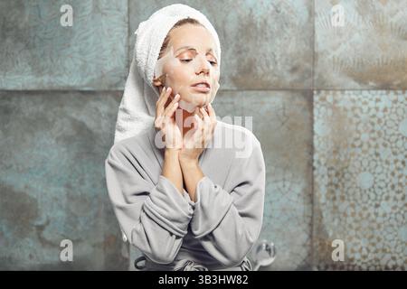 Erwachsenes Mädchen mit einer Papier-Spa-Maske auf dem Gesicht, sitzt im Badezimmer, trägt einen Bademantel und ein Handtuch auf dem Kopf. Stockfoto