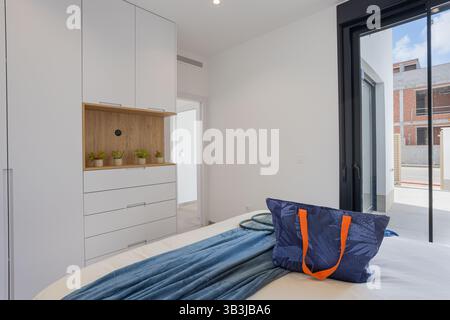 Geräumiges Schlafzimmer mit sauberen weißen Wänden, eingebautem Kleiderschrank, blauer Tasche auf dem Bett und Zugang zur privaten Terrasse durch Glastüren. Stockfoto