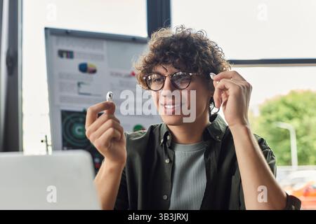 Ein junger Profi lächelt bei der Arbeit, trägt kabellose Ohrstöpsel und ist umgeben von Büroartikeln. Stockfoto