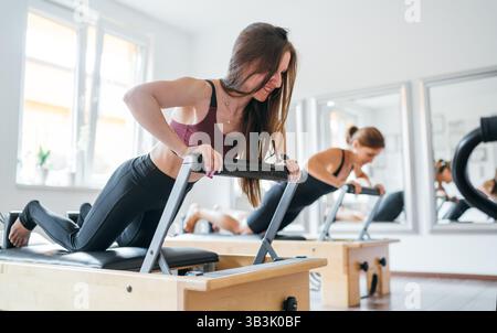 Two young females doing extended plank static strengthening core muscles exercise using pilates reformer machine in sport athletic gym hall. Active pe Stockfoto
