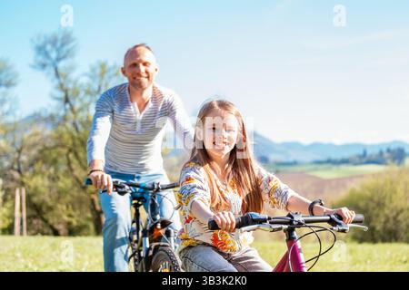 Porträt eines lächelnden Vaters mit seiner Tochter während einer Fahrradtour im Freien. Sie genießen eine gute gemeinsame Zeit in einem sonnigen Stadtpark. Glückliche Par Stockfoto