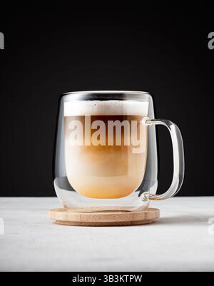 Eleganter Cappuccino-Becher in transparentem Glas auf Holzuntersetzer, Lifestyle-Themen Stockfoto