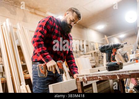 Geschulter Zimmermann, der in einer Werkstatt mit einem Bohrschrauber Holzmöbel baut Stockfoto