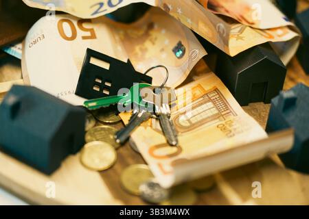 Hausschlüssel in Nahaufnahme mit kleinem Schlüsselanhänger in Form eines Hauses, der auf Euro-Banknoten und Münzen platziert ist. Umschlossene Miniaturhaushäuser, die die Kaufkosten pro Jahr symbolisieren Stockfoto