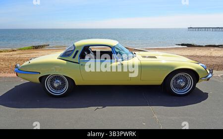Klassischer gelber Jaguar XKE V12 Roadster, der an der Strandpromenade am Strand und am Meer im Hintergrund geparkt ist Stockfoto