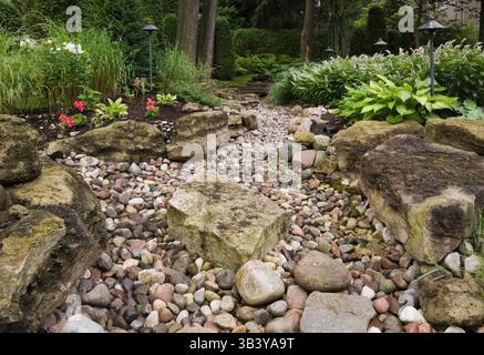 Trockener Bach begrenzt von rotem Pelargonium - Geranien, Miscanthus - Ziergras, Hosta und Lysimachia clethroides - Loosestrife im Garten. Stockfoto