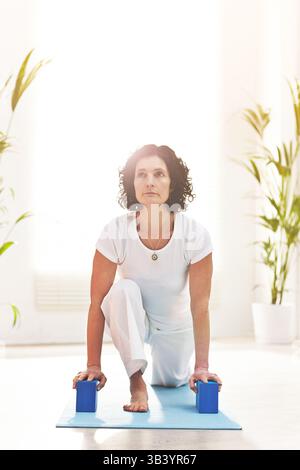 Reife Frau, die Yoga übt, während sie sich in einem hellen Studio auf zwei Schaumstoffblöcke lehnt. Yogi benutzt Ausrüstung, während er allein zu Hause trainiert. Aktive Dame Stockfoto