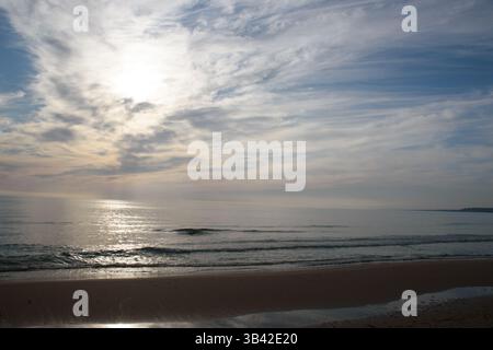Sonnenstrahlen brechen durch schwere Wolken über einem ruhigen Meereshorizont und erzeugen eine dramatische und kraftvolle natürliche Lichtshow während eines stimmungsvollen Sonnenuntergangs an der Küste. Stockfoto