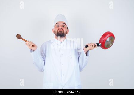 Porträt eines jungen männlichen Küchenchefs mit Pfanne und Holzlöffel in weißer Uniform und verwirrter Vorderansicht Stockfoto
