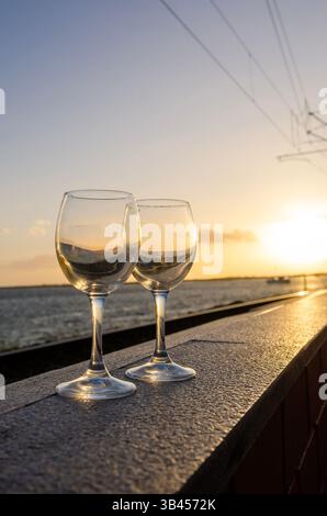 Leere Weingläser stehen auf einem Felsvorsprung mit Blick auf das ruhige Wasser und spiegeln die warmen Farbtöne eines malerischen Sonnenuntergangs wider Stockfoto