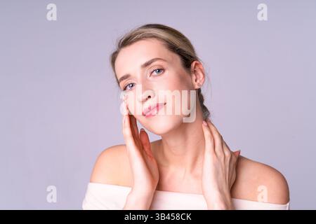 Junge Frau mit blauen Augen, die sanft ihr Gesicht berührt und auf hellem Hintergrund Pflege für ihre Haut zeigt. Stockfoto
