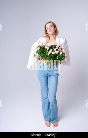 Eine junge Frau, die einen großen Rosenstrauß in blauer Jeans und einem weißen Hemd barfuß in einem hellen Studio hält. Geeignet für Freude, Feiern und Stolz Stockfoto