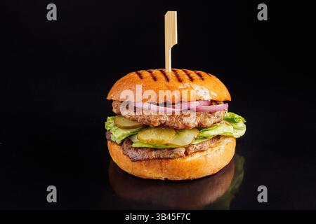 Saftiger gegrillter Rindfleischburger mit Gurken, Salat, Zwiebeln in einem gerösteten Brötchen auf schwarzem Hintergrund, Nahaufnahme. Stockfoto