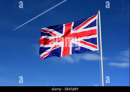 Die britische Flagge flattert im Wind ein Flugzeug fliegt in großer Höhe mit einem Kondensstreifen vorbei Stockfoto