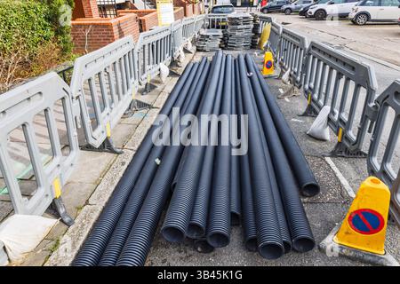 Gestapelte schwarze Kunststoffrohre auf der städtischen Straßenbaustelle mit Sicherheitsbarrieren und Kegeln Stockfoto