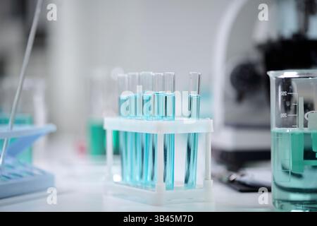 Laborausrüstung mit mehreren Reagenzgläsern, die mit einer leuchtend blauen Flüssigkeit gefüllt sind. Im Hintergrund ist ein Becher mit einer Pipette zu sehen, was auf eine wissenschaftliche Versuchsumgebung hinweist Stockfoto