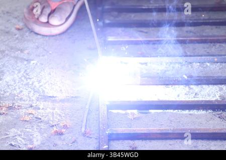 Man schweißt Metallrahmen außen mit leuchtenden Funken. Asiatischer Mann wird in Sandalen gezeigt. Schweißen erzeugt helles Licht und Rauch im Außenbereich. Stockfoto