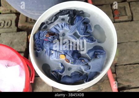 Top-Down-Shot erfasst Jeans aus blauem Denim, die in einem weißen Kunststoffeimer in Schaumwasser getaucht sind und für eine gründliche Reinigung im Freien bereit sind. Stockfoto