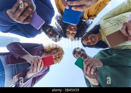 Gruppe fröhlicher junger Schüler, die Smartphones benutzen, im Kreis vor blauem Himmel stehen, aus tiefem Blickwinkel Stockfoto