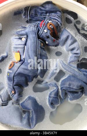 Blaue Jeansjacke aus Denim im in Seifenwasser gefülltem Becken. Kleidung mit der Hand waschen, Denimtuch in schaumigem Wasser einweichen, Körperpflege, Stoffkonzept. Stockfoto