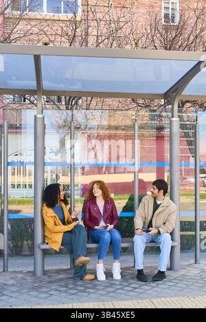 Gruppe verschiedener junger Studenten, die ihre Smartphones benutzen und miteinander plaudern, während sie an der Bushaltestelle auf den Bus warten Stockfoto