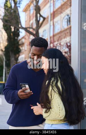 Junge Mann und Frau stehen zusammen auf einer belebten Straße, lächeln, während sie ihre Smartphones durchsuchen und den Moment genießen Stockfoto
