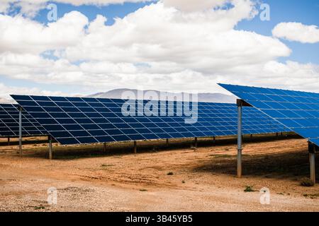 Mehrere Reihen blauer Photovoltaik-Solarpaneele in einem wüstenartigen offenen Feld mit trockenem Boden und Bergkulisse installiert Stockfoto