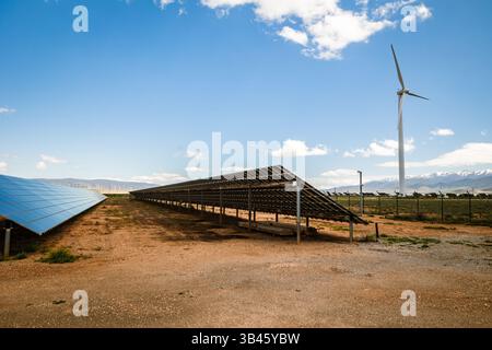 Eine Hybrid-Anlage für erneuerbare Energien mit Solarpaneelen neben einer großen Windturbine. Auf trockenem Boden mit klarem, blauem Himmel Stockfoto