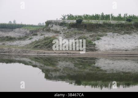 Scenic Shot zeigt eine erodierte Erdklippe, die von karger Vegetation bedeckt ist. Ein stiller Fluss reflektiert die Landschaft und schafft spiegelnde Bilder von Grün Stockfoto