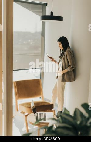 Eine junge Frau lehnt sich an eine Wand in einem eleganten Arbeitsbereich, eingetaucht in ihr Telefon. Stockfoto