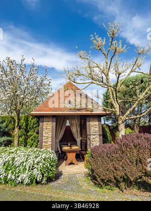 Schöne Hinterhof-Landschaftsgestaltung mit einem steinernen Pavillon, blühenden Bäumen und Büschen im Frühling. Eleganter Gartentavillon mit Ziegeldach inmitten blühender Stockfoto