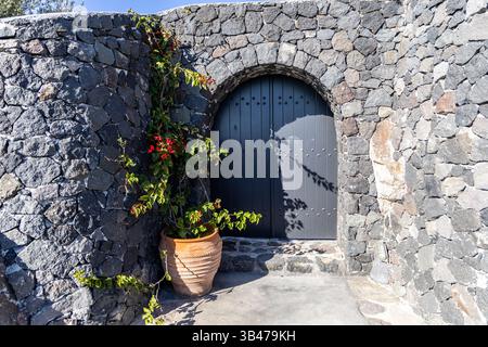 Eingang mit dunkler Holztür, eingerahmt von Steinmauer und Bougainvillea in Santorin. Stockfoto